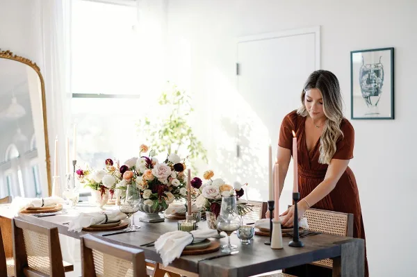 A woman in a rust-colored dress arranging a wedding table setting with floral centerpieces and candles.