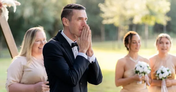 A groom in a black tuxedo covers his mouth with his hands during an outdoor wedding ceremony, with bridesmaids in the background.