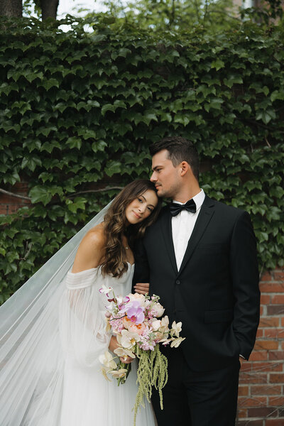 A bride and groom posing together in front of a leafy wall, with the bride holding a bouquet.