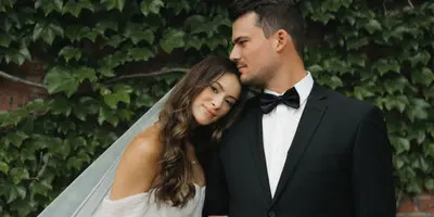 A bride and groom posing together in front of a leafy wall, with the bride holding a bouquet.