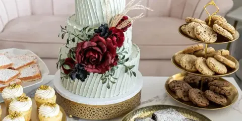 A wedding dessert table featuring a tiered white cake with floral decorations, a stand of cupcakes, a tiered stand of cookies, and a plate of brownies.