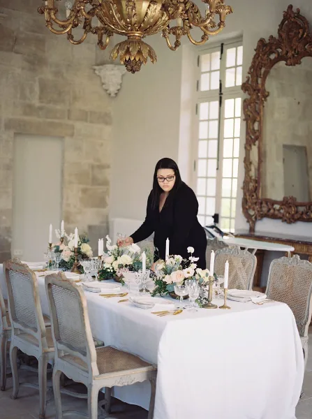 A professional event designer arranging a floral centerpiece on a long banquet table in a historic venue with a gilded chandelier.