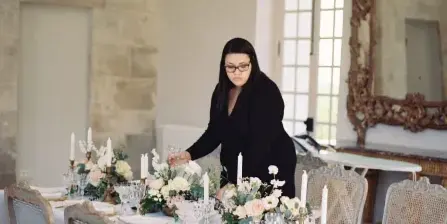 A professional event designer arranging a floral centerpiece on a long banquet table in a historic venue with a gilded chandelier.