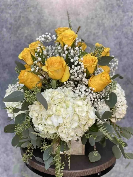 A floral arrangement featuring yellow roses, white hydrangeas, and eucalyptus leaves arranged in a vase on a wooden stand.
