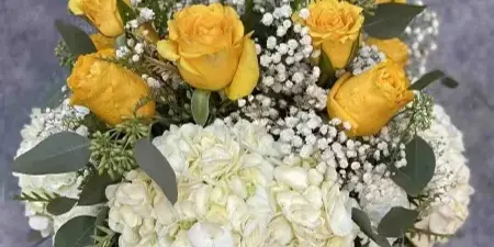 A floral arrangement featuring yellow roses, white hydrangeas, and eucalyptus leaves arranged in a vase on a wooden stand.