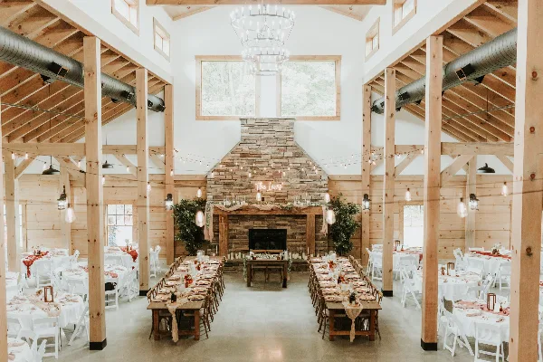 Interior view of a rustic wedding venue with exposed wooden beams, a large stone fireplace, and long dining tables set for a reception.