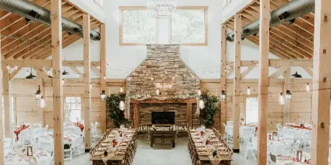 Interior view of a rustic wedding venue with exposed wooden beams, a large stone fireplace, and long dining tables set for a reception.