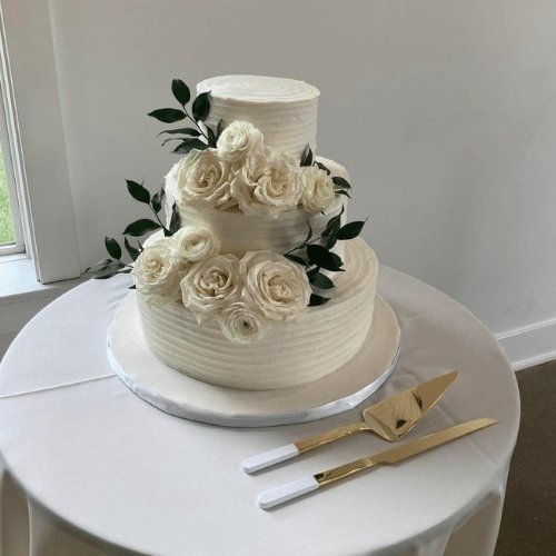 A three-tiered white wedding cake with buttercream roses and greenery on a table with gold cake servers.