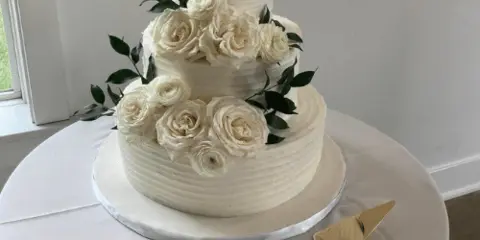 A three-tiered white wedding cake with buttercream roses and greenery on a table with gold cake servers.