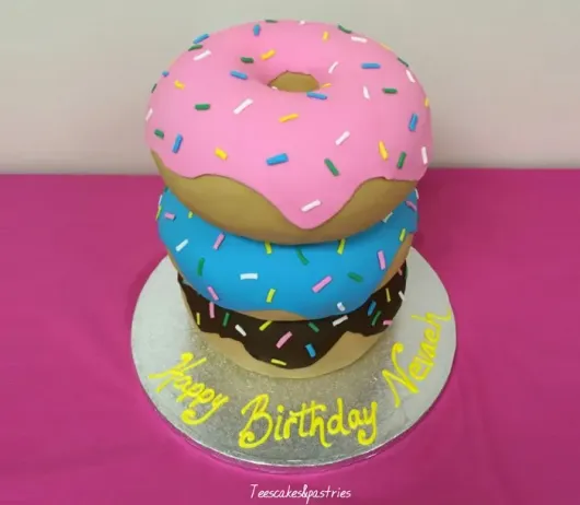 A three-tiered specialty cake designed to look like stacked donuts with pink, blue, and chocolate frosting and sprinkles, sitting on a silver cake board with 'Happy Birthday' written in yellow icing.