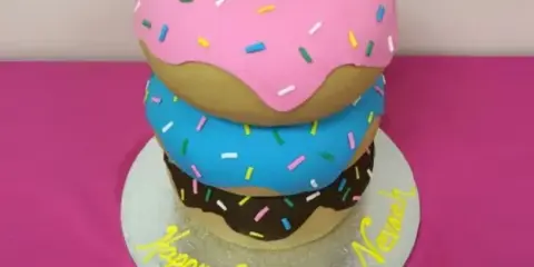 A three-tiered specialty cake designed to look like stacked donuts with pink, blue, and chocolate frosting and sprinkles, sitting on a silver cake board with 'Happy Birthday' written in yellow icing.