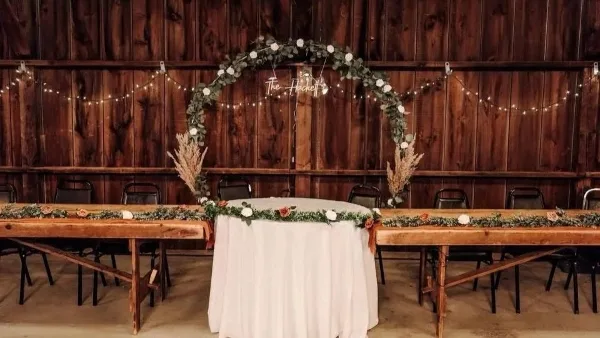 A wedding head table setup with a floral arch and string lights against a dark wooden barn wall.