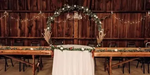 A wedding head table setup with a floral arch and string lights against a dark wooden barn wall.