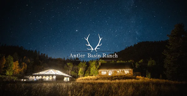 A night view of a rustic log cabin and a large white event tent on a grassy field under a starry sky.