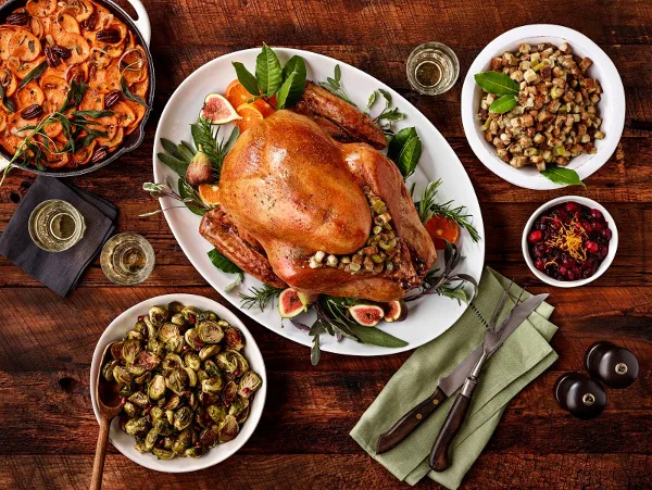 A top-down view of a gourmet catering spread featuring a roasted turkey, roasted Brussels sprouts, stuffing, and a tomato dish on a wooden table.