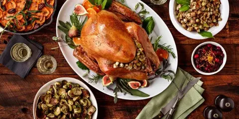 A top-down view of a gourmet catering spread featuring a roasted turkey, roasted Brussels sprouts, stuffing, and a tomato dish on a wooden table.