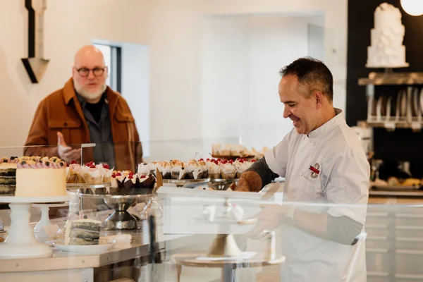 A baker in a white uniform smiles while serving a customer at a bakery counter filled with cakes and pastries.
