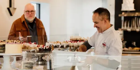 A baker in a white uniform smiles while serving a customer at a bakery counter filled with cakes and pastries.
