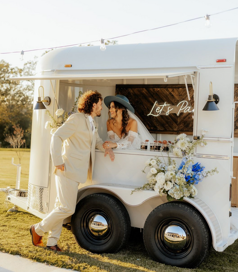 A bride and groom posing at a white mobile bar trailer rental decorated with flowers and string lights.