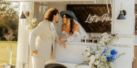 A bride and groom posing at a white mobile bar trailer rental decorated with flowers and string lights.