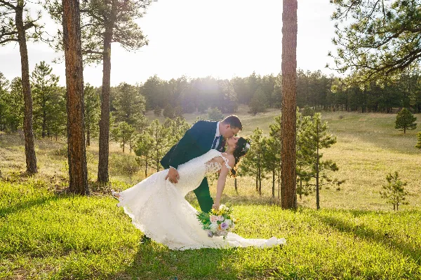 A bride and groom sharing a kiss in a grassy field surrounded by pine trees.