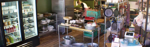 Interior view of a commercial kitchen with a chef preparing food, refrigerated display cases, and shelves stocked with prepared goods and kitchen equipment.