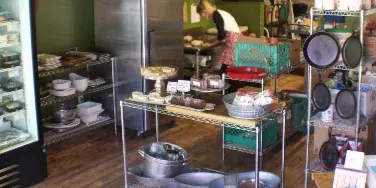 Interior view of a commercial kitchen with a chef preparing food, refrigerated display cases, and shelves stocked with prepared goods and kitchen equipment.