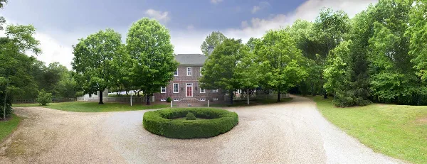 A colonial-style brick building with a red door, surrounded by green trees and a circular hedge in a gravel driveway.