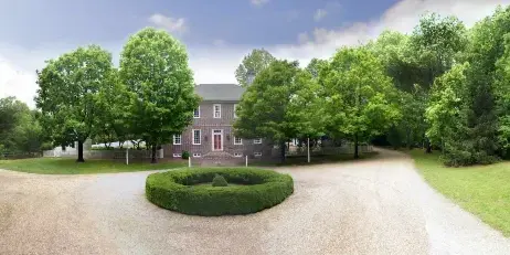 A colonial-style brick building with a red door, surrounded by green trees and a circular hedge in a gravel driveway.