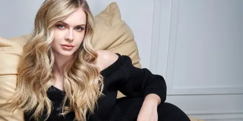 Portrait of a woman with long, styled blonde wavy hair seated on a tan leather chair.