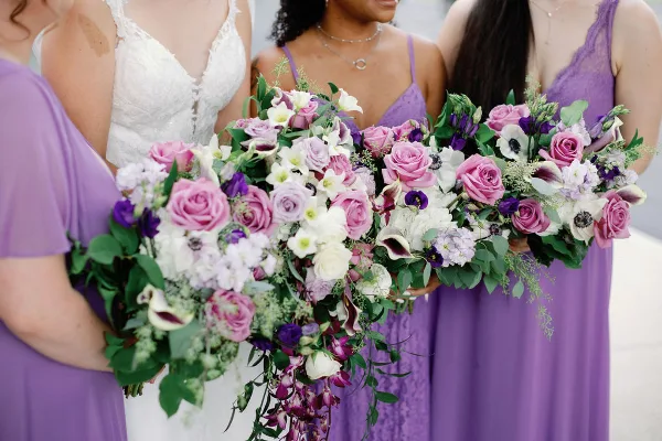 Close-up of a bridal bouquet and bridesmaids' bouquets featuring pink roses, white calla lilies, and purple accents.