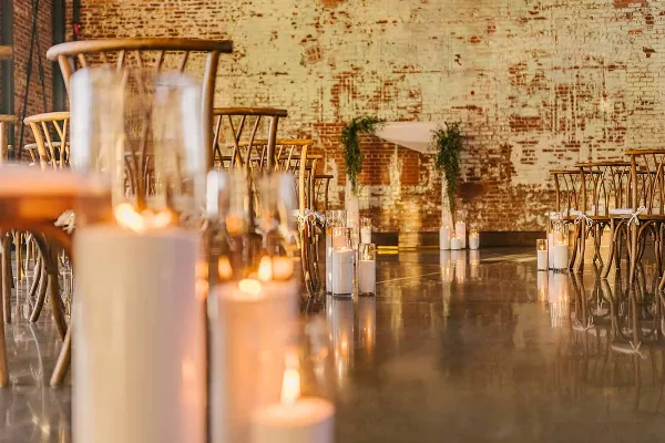 A wide shot of a wedding venue with a polished floor, featuring a large arrangement of lit pillar candles and hurricane glass holders set up for a ceremony or reception.