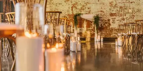 A wide shot of a wedding venue with a polished floor, featuring a large arrangement of lit pillar candles and hurricane glass holders set up for a ceremony or reception.