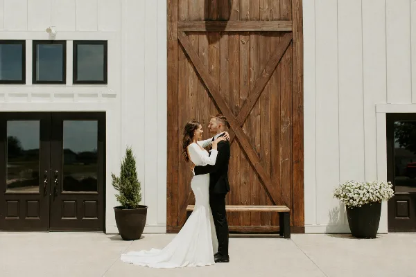 A bride and groom embracing in front of a modern white barn with a large wooden door.