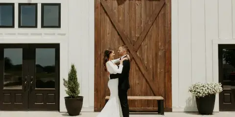 A bride and groom embracing in front of a modern white barn with a large wooden door.
