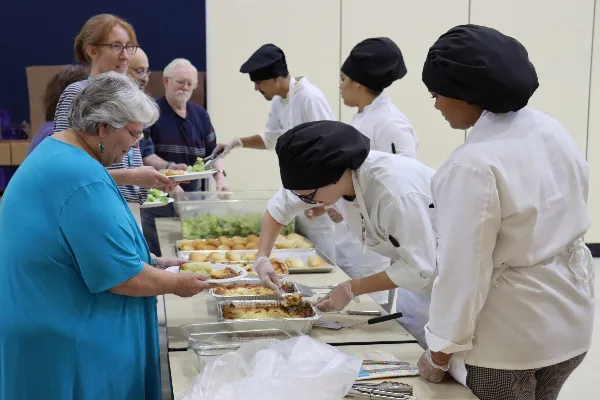 Culinary students in white chef coats and black hats serving lasagna and rolls to older adults at a buffet table.