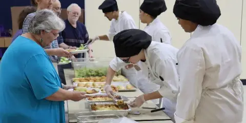 Culinary students in white chef coats and black hats serving lasagna and rolls to older adults at a buffet table.