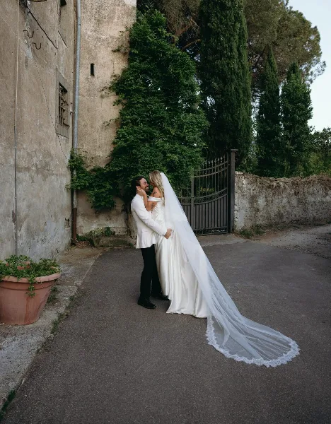 A bride and groom embracing in a rustic outdoor setting with a long veil.