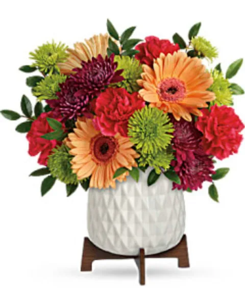 A vibrant floral arrangement featuring orange gerbera daisies, red and purple chrysanthemums, and green button mums in a white textured vase.