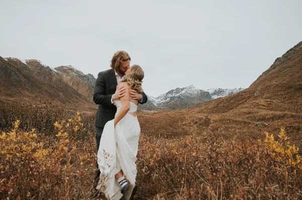 A bride and groom embracing in a mountainous field with snow-capped peaks in the background.