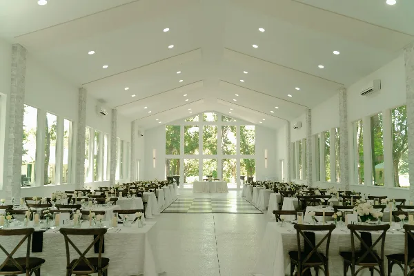 Interior view of a white glasshouse wedding reception hall with stone pillars, set tables, and large windows looking out onto trees.