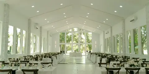 Interior view of a white glasshouse wedding reception hall with stone pillars, set tables, and large windows looking out onto trees.