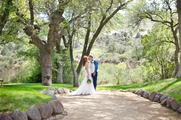 A bride and groom kissing outdoors in a grassy area with trees and a dirt path.