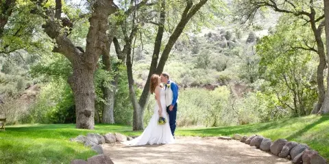 A bride and groom kissing outdoors in a grassy area with trees and a dirt path.