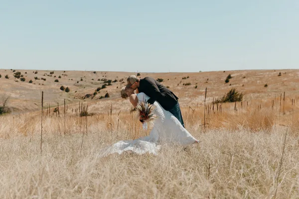 A bride and groom kissing in a field of dry golden grass under a clear blue sky.
