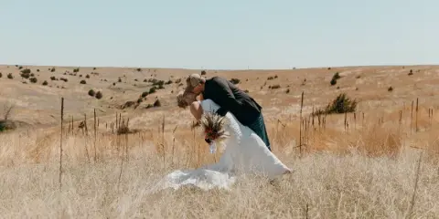 A bride and groom kissing in a field of dry golden grass under a clear blue sky.