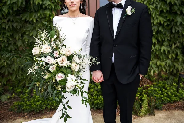 A bride and groom standing together, with the bride holding a large bouquet of white and blush roses and greenery.