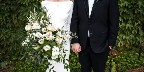 A bride and groom standing together, with the bride holding a large bouquet of white and blush roses and greenery.