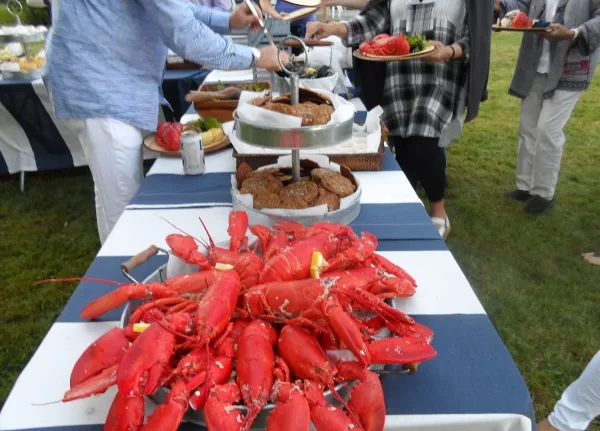 An outdoor catering event featuring a large table laden with cooked lobsters and other food, with guests serving themselves.