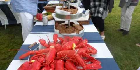An outdoor catering event featuring a large table laden with cooked lobsters and other food, with guests serving themselves.